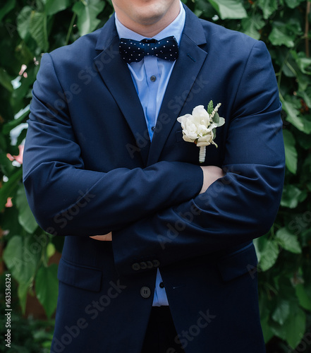 Stylish groom in blue suit, bowtie and boutonniere standing with crossed hands. Wedding day