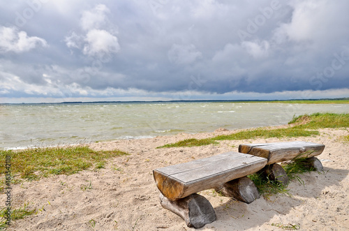 Empty beach on the lake with rustic wooden benches, stormy clouds. Wide angle view.