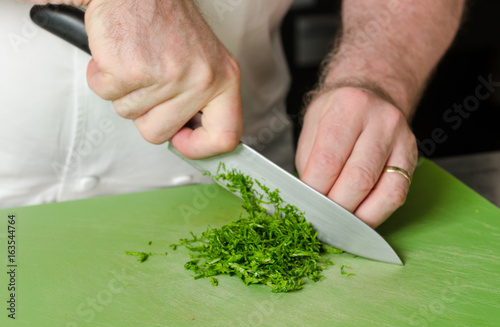 A knife, being held by male hands, slicing through fresh organic parsley, on a green vegetable chopping board