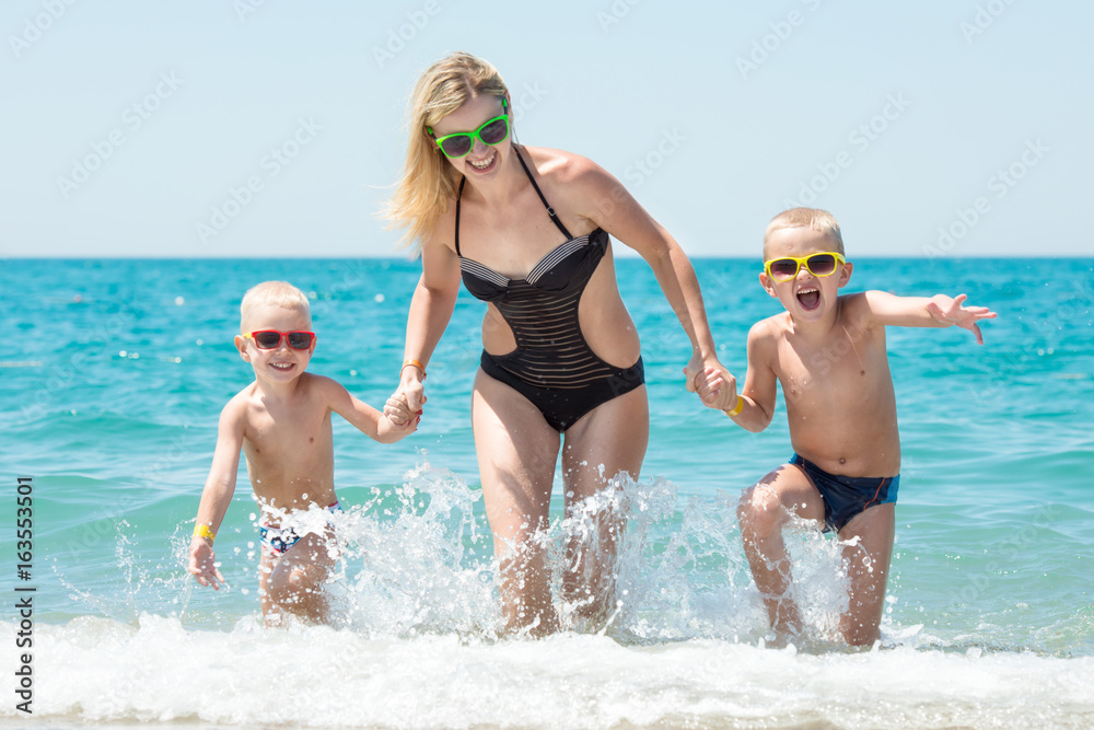 Beautiful mother and two lovely son holding hands running on the waves.Fun,family-friendly summer vacation.