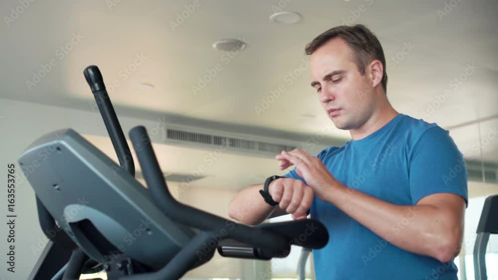 Handsome fit young man in gym doing exercises on exercise bike. Using his smartwatch checking vitals Handsome fit young man in gym doing exercises on exercise bike. Using his smartwatch checking vitals
