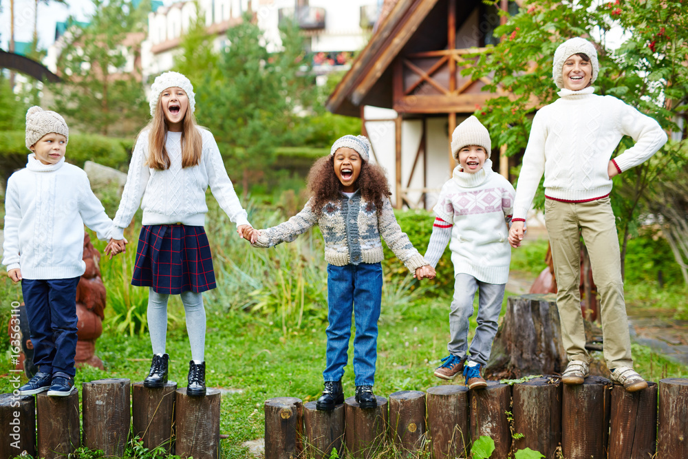 Happy children of various ages having fun playing outdoors in green countryside, standing in row on wooden posts holding hands dressed in similar white knit clothes