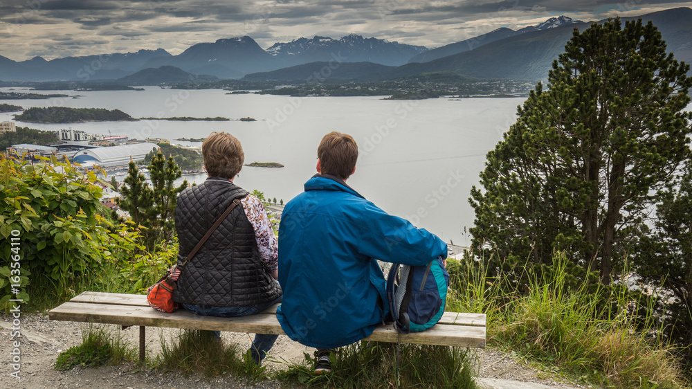 Fototapeta premium A man and a woman are sitting on a bench and looking at the Norwegian panorama. Theme of tourism