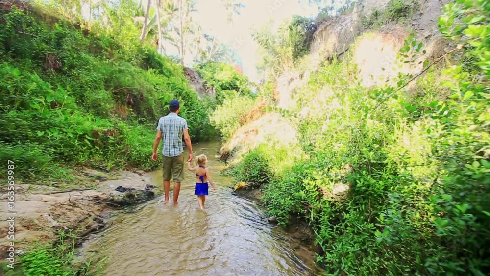 Dad little daughter in blue swimsuit walk barefoot along narrow Fairy stream between green bush bank and steep rock bank Dad little daughter in blue swimsuit walk barefoot along narrow Fairy stream between green bush bank and steep rock bank