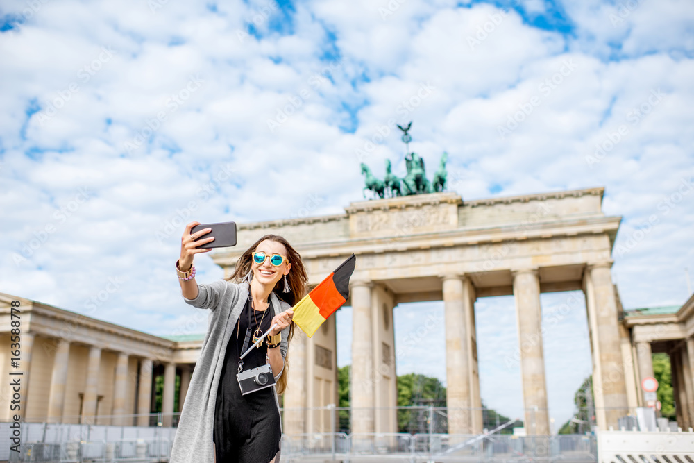 Fototapeta premium Young woman tourist making selfie photo with german flag standing in front of the famous Brandenburg gates in Berlin