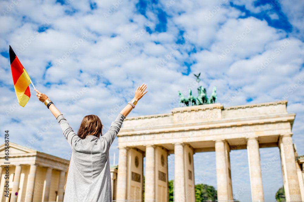 Obraz premium Młoda kobieta turysta stojąc z podniesionymi rękami i niemiecką flagą przed słynną bramą brandenburską w Berlinie