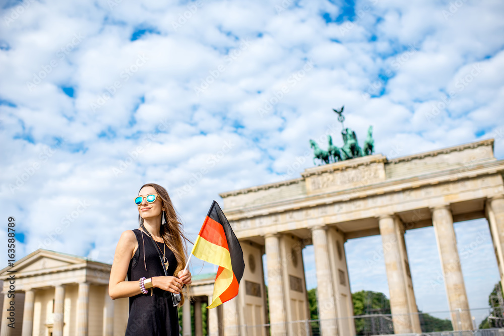 Fototapeta premium Portrait of a young smiling woman tourist standing with german flag in front of the famous Brandenburg gates in Berlin