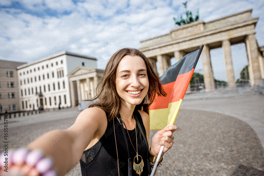 Fototapeta premium Young woman tourist making selfie photo with german flag standing in front of the famous Brandenburg gates in Berlin