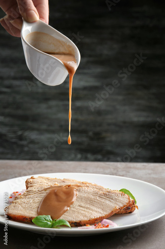 Pouring tasty turkey gravy onto sliced meat on plate against dark background