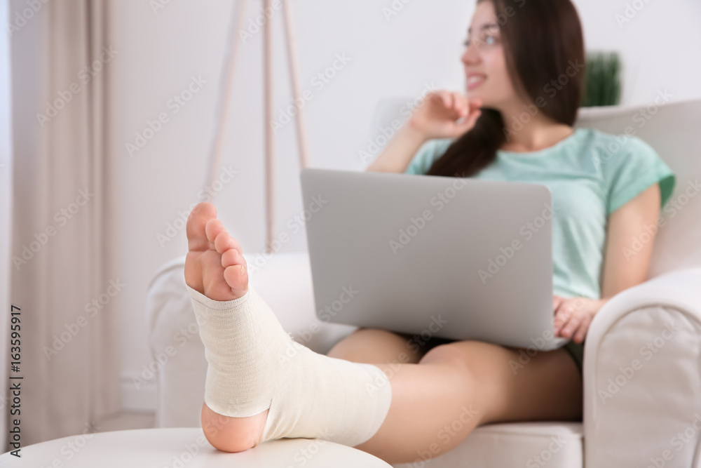 Fototapeta premium Beautiful young woman with laptop and bandaged leg sitting in arm-chair at home