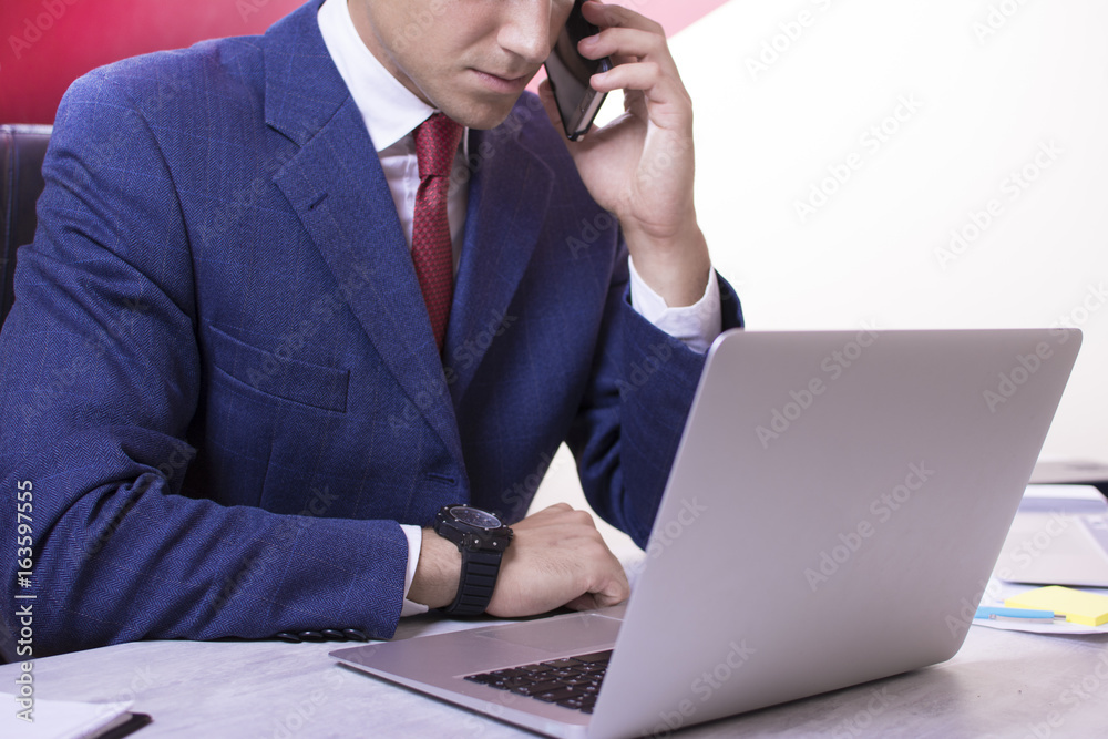 Close up view of a young businessman working on his desk white background. Two computers on the table. Man making a phone call while looking at laptop screens. Red tie and blue suit. Close up view of a young businessman working on his desk white background. Two computers on the table. Man making a phone call while looking at laptop screens. Red tie and blue suit.