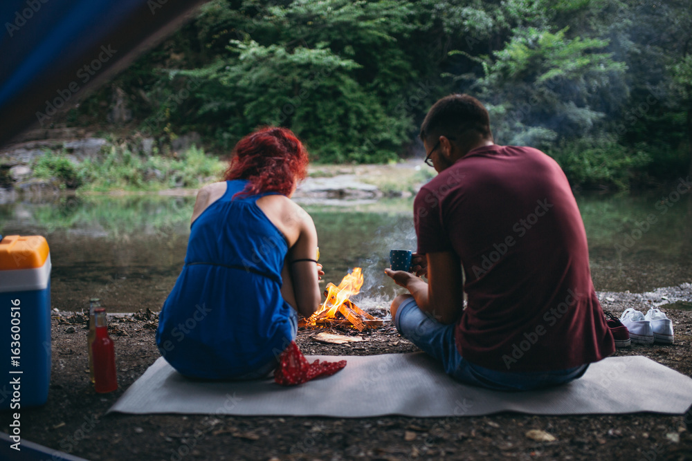 Happy young couple camping and hiking on wild mountain river. They sitting around campfire and drinking coffee. View from the tent.