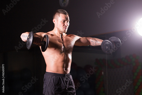 Athletic shirtless young male fitness model holds the dumbbells.
