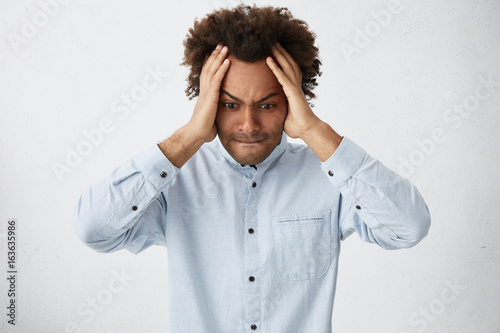 Young dark-skinned hipster guy wearing white shirt holding hands on head looking desperately down going to cry while found out about something horrorful. Crying man with bushy hair having troubles photo
