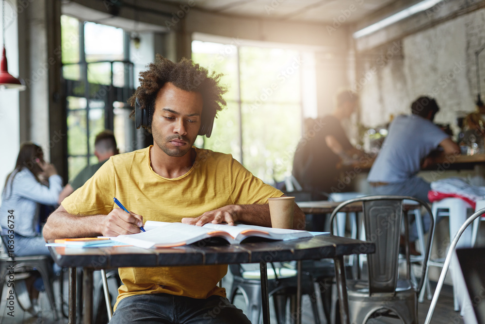 People, youth and education concept. Carefree student dressed in yellow T-shirt preparing for classes at university while sitting at desk in cafe listening to music with headphones having serious look
