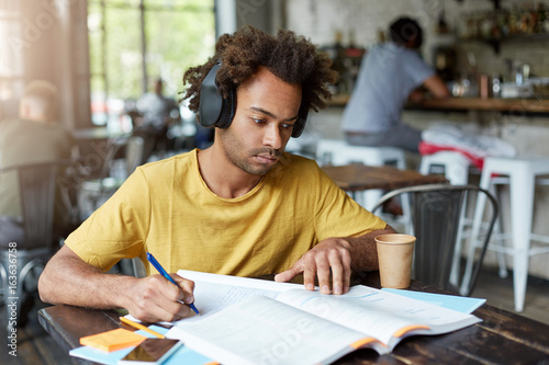 Hipster Afro American guy doing his home assigment looking attentively in book and copybook listening to music in headphones and drinking coffee while sitting at cozy restaurant. Education concept