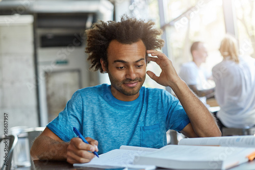 Portrait of handsome African American male with bushy hair sitting at desk in university canteen writing notes scratching his head not knowing something prepearing scientific research or project