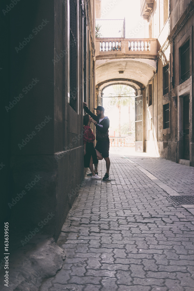 A bearded man in a cap wants to kiss his girlfriend while standing beside the building wall on a side street. Beautiful woman is looking with love at her beloved man during romantic dating.