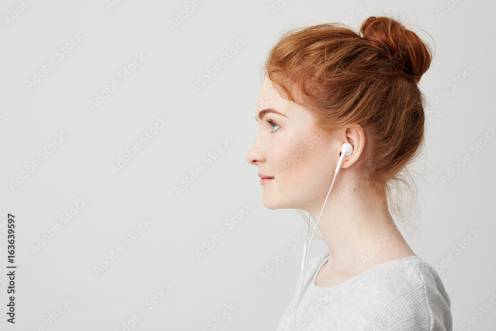 Profile portrait of young beautiful tender redhead girl with bun in headphones smiling over white background. Profile portrait of young beautiful tender redhead girl with bun in headphones smiling over white background.