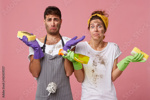 Portrait of unhappy male and female with dirty faces and clothes holding washing spray and sponges shrugging shoulders being sad not knowing how to remove all stains on windows. Facial expressions