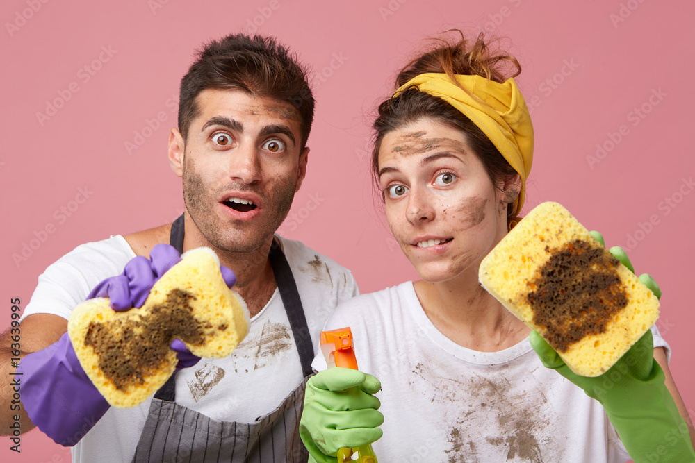 Φωτογραφία Astonished female and her husband with dirty faces showing very dirty sponges after cleaning window sill posing against pink background