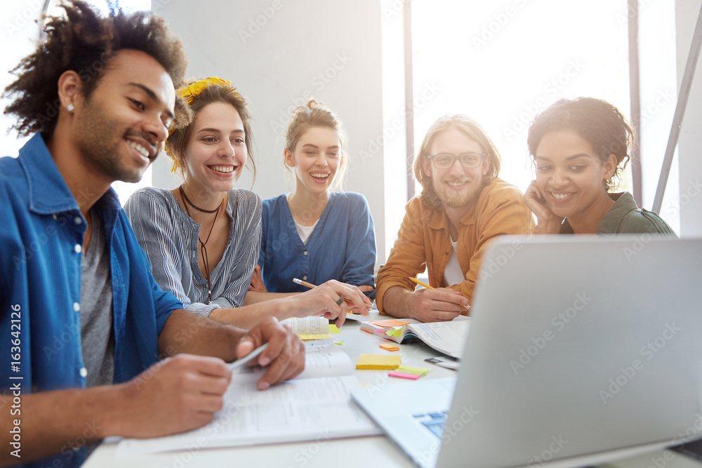Obraz premium Group of five mixed race students sitting together surrounded with books looking happily at laptop making video call to their friend abroad being glad to see him. University friends with books, laptop