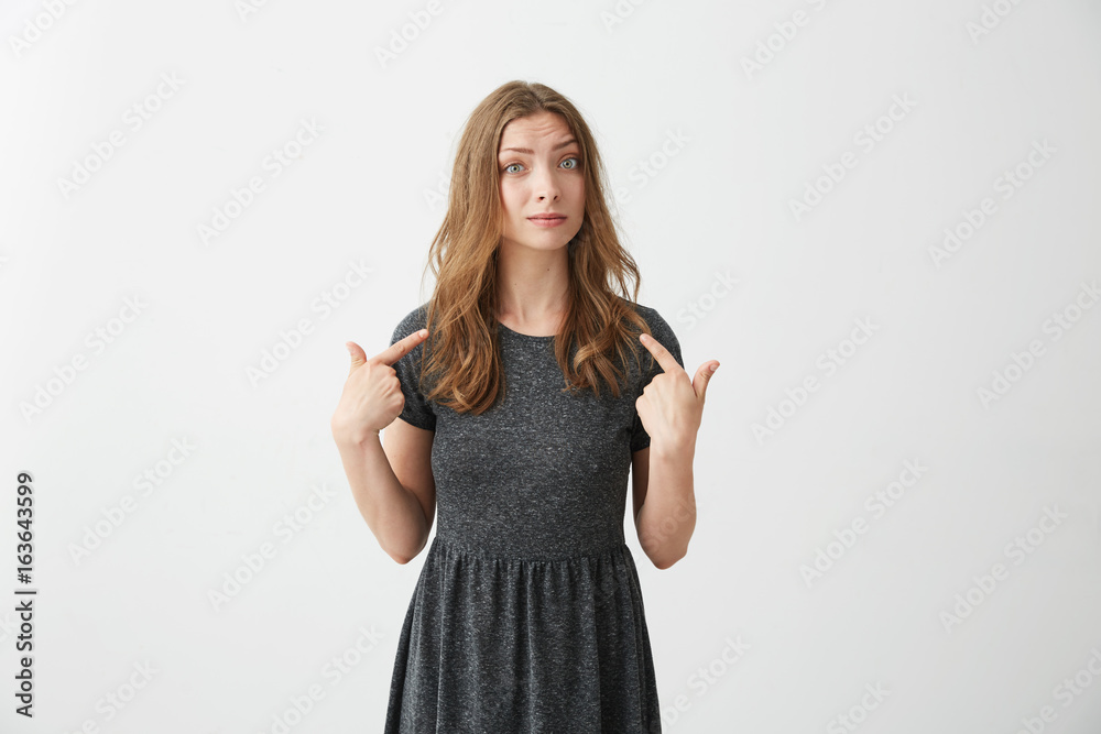 Portrait of surprised young beautiful girl looking at camera pointing fingers at herself over white background. Portrait of surprised young beautiful girl looking at camera pointing fingers at herself over white background.