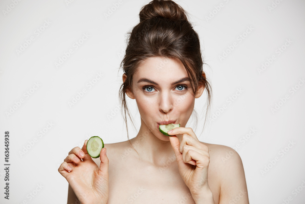 Pretty young natural girl with perfect clean skin looking at camera eating cucumber slice over white background. Facial treatment. Pretty young natural girl with perfect clean skin looking at camera eating cucumber slice over white background. Facial treatment.