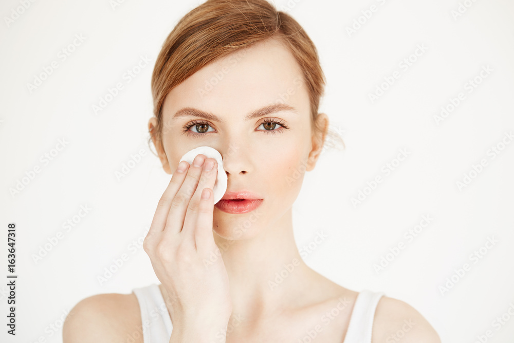 Beautiful natural blonde girl cleaning face with cotton sponge looking at camera over white background. Cosmetology and spa. Beautiful natural blonde girl cleaning face with cotton sponge looking at camera over white background. Cosmetology and spa.