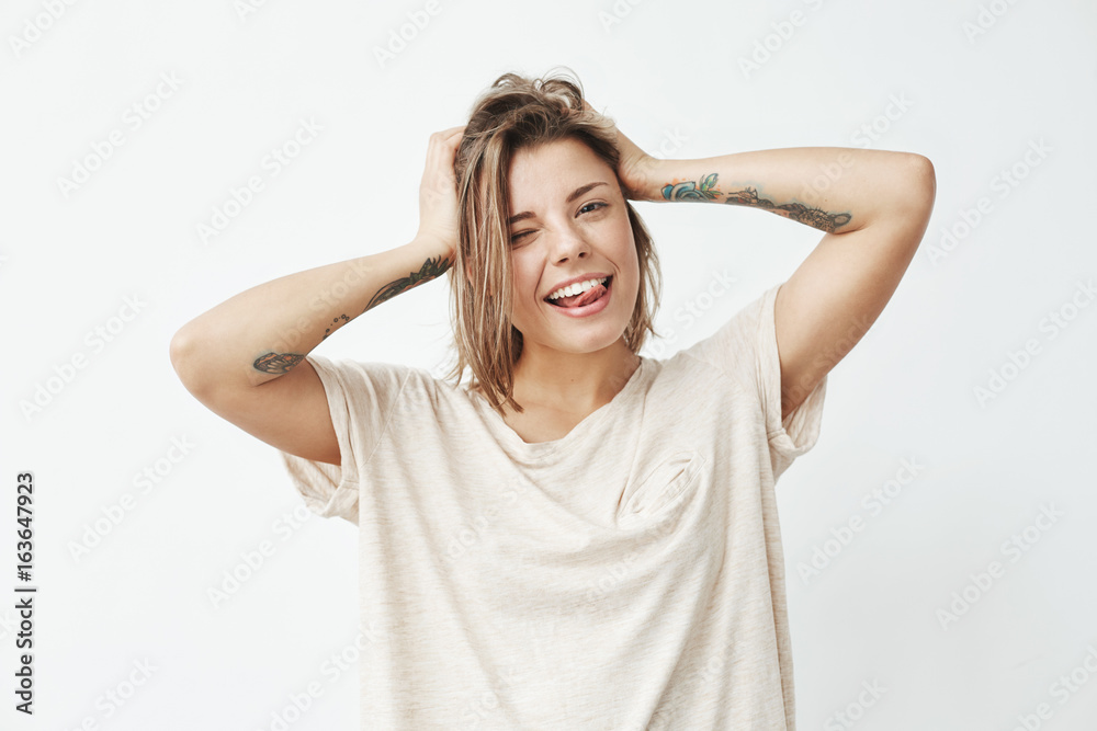 Cheerful cool tattooed girl smiling winking showing tongue looking at camera touching hair over white background. Cheerful cool tattooed girl smiling winking showing tongue looking at camera touching hair over white background.