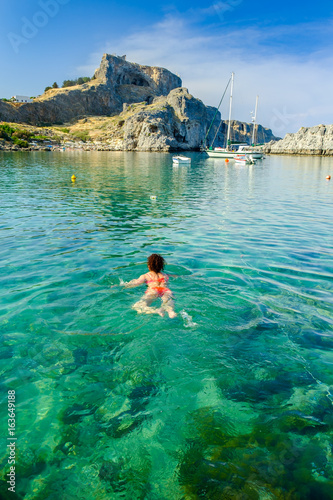 Woman swimming in St.Paul's Bay with Lindos Castle and the Acropolis at the background.