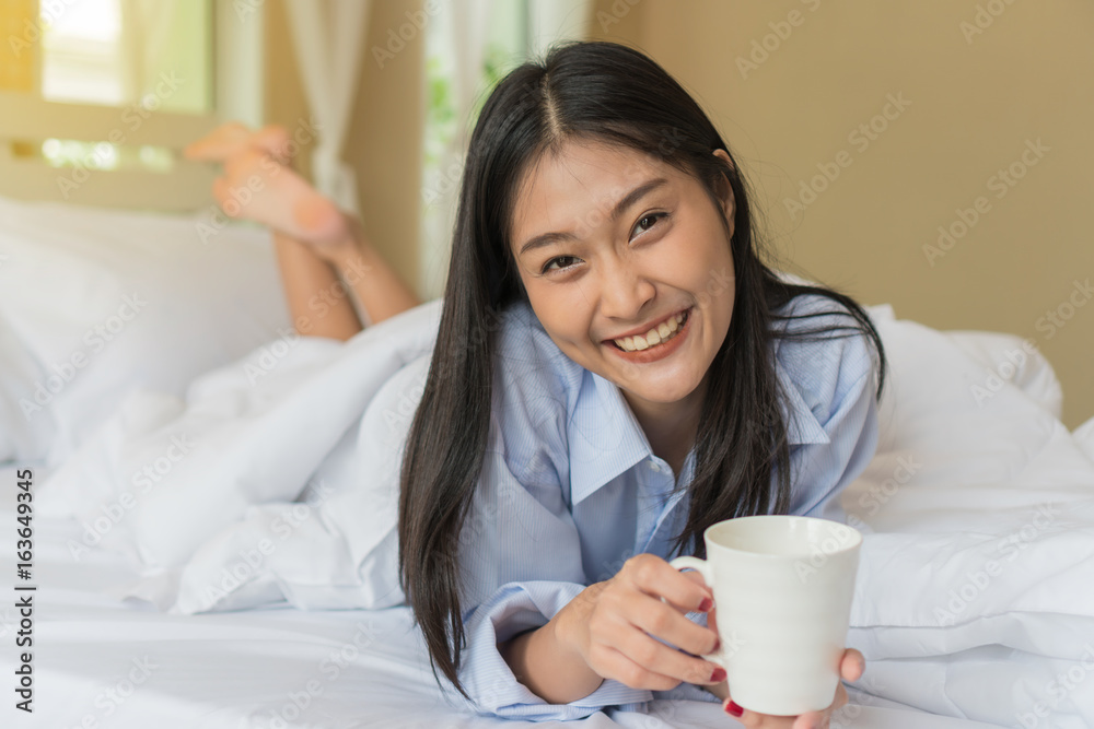 Happy smiling Asian young woman lying holding a cup of coffee on bed. Happy smiling Asian young woman lying holding a cup of coffee on bed.