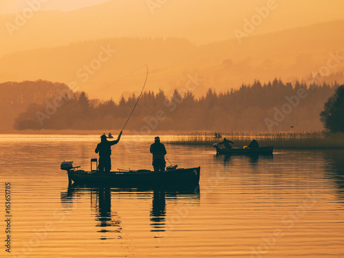 Silhouette of man fishing on lake from boat at sunset. Lake of Menteith, Stirlingshire, Scotland, UK