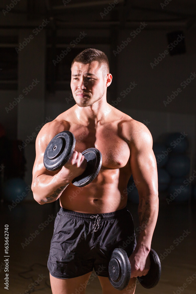 Handsome power athletic guy bodybuilder doing exercises with dumbbell. Fitness muscular body on dark background