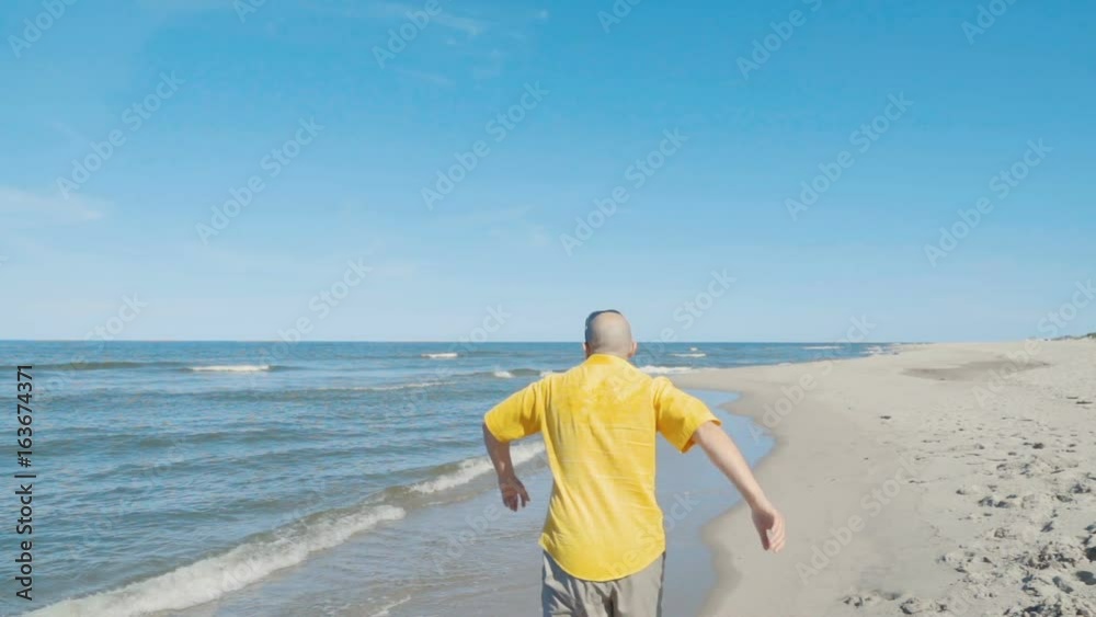 Young bald man in a yellow shirt runs and jumps into the water barefoot and enjoys herself. Beach with white sand. Outgoing coastal line. Slow Motion.