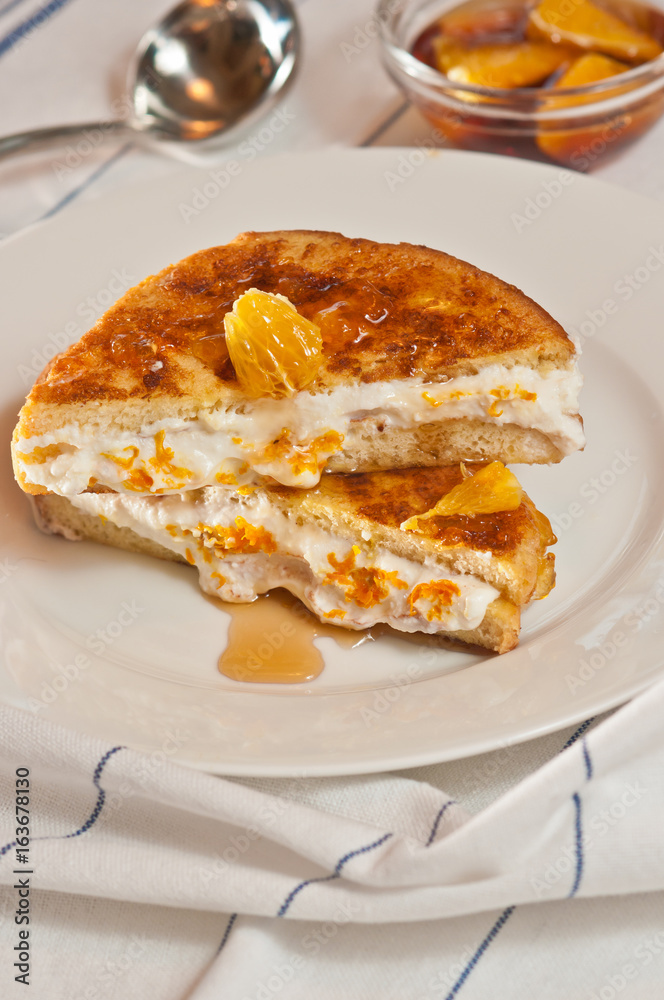 Orange cream french toast sandwich on a round, white plate and bowl of orange sections and an artisan spoon on a white with blue striped cloth Orange cream french toast sandwich on a round, white plate and bowl of orange sections and an artisan spoon on a white with blue striped cloth