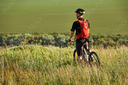 Rear view of the sportsman with his mountain bike stands on the meadow and looking away in the countryside.