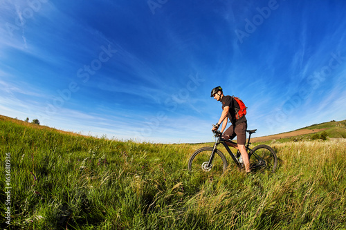 Young cyclist cycling on the green meadow against beautiful blue sky with clouds in the countryside.
