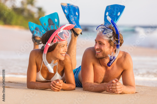 Summer travel beach vacation snorkel couple talking together laughing having fun relaxing lying down on sand at sunset. Girl, man wearing diving mask and fins happy.