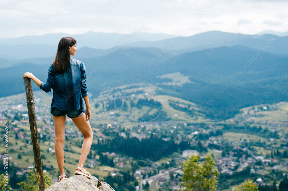 Naklejka premium Lonely beautiful long haired young traveler girl with sexy legs in jeans clothes standing on stone at top of carpathian mountain with wooden staff stick and looking at village far at horizon.