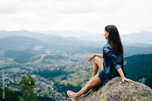 Lonely young traveler girl siiting on stone at top of mountain. Discovering new country. Beautiful nature landscape from high altitude. Summer trip on weekend vacation. Edge of world. Jeans clothes.