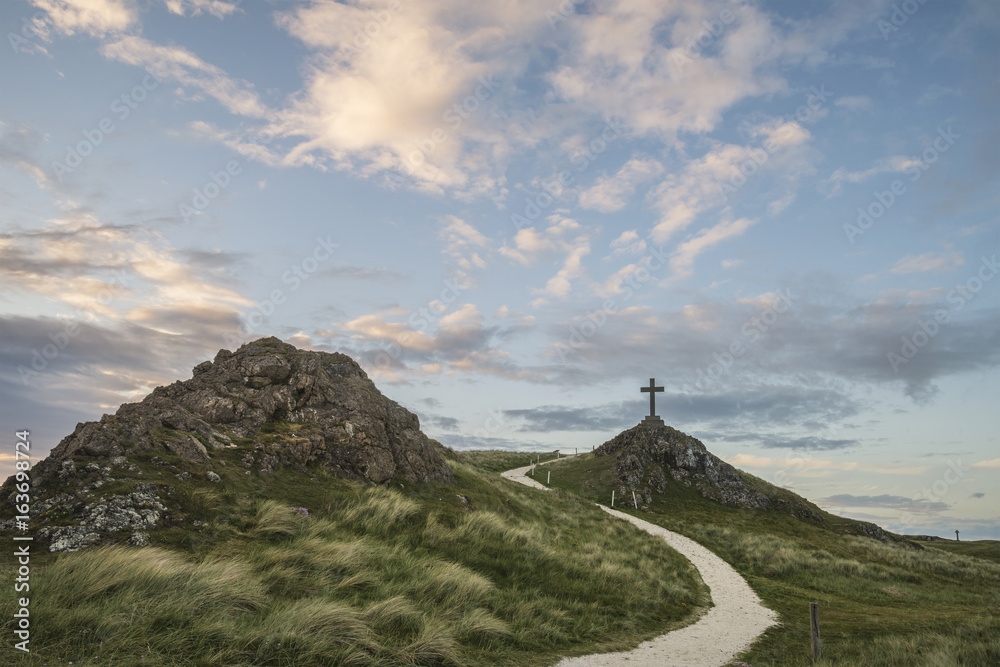 Cross in landscape of Ynys Llanddwyn Island with Twr Mawr lighthouse in background with blue sky