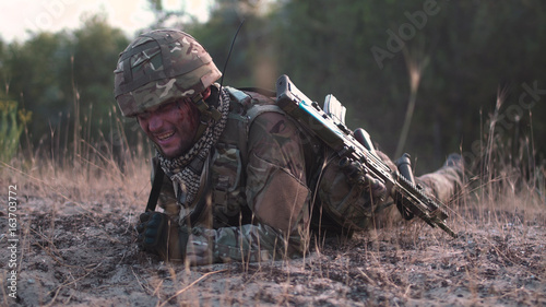 Man in military uniform with gun crawling alone on ground in field.