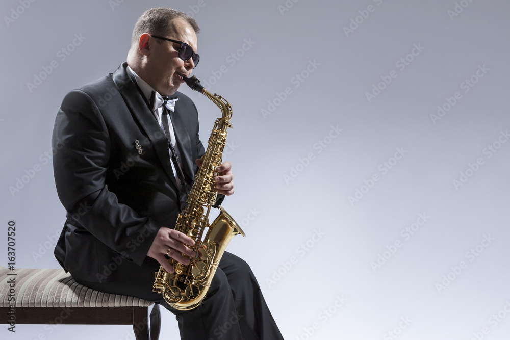 Fototapeta premium Music Concepts. Portrait of Mature Relaxed and Thoughful Caucasian Saxophone Player in Sunglasses Playing the Saxophone While Sitting on Chair in Studio Environment.