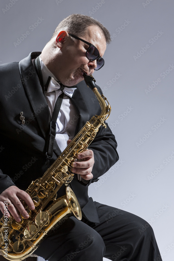 Fototapeta premium Male Mature Saxophone Player in Sunglasses Playing the Saxophone While Sitting on Chair in Studio Environment.