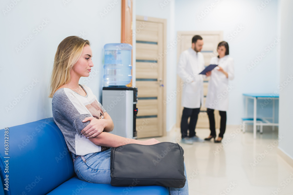 Young woman filling nervous sitting at hospital corridor waiting while two doctors talking on a backgroud