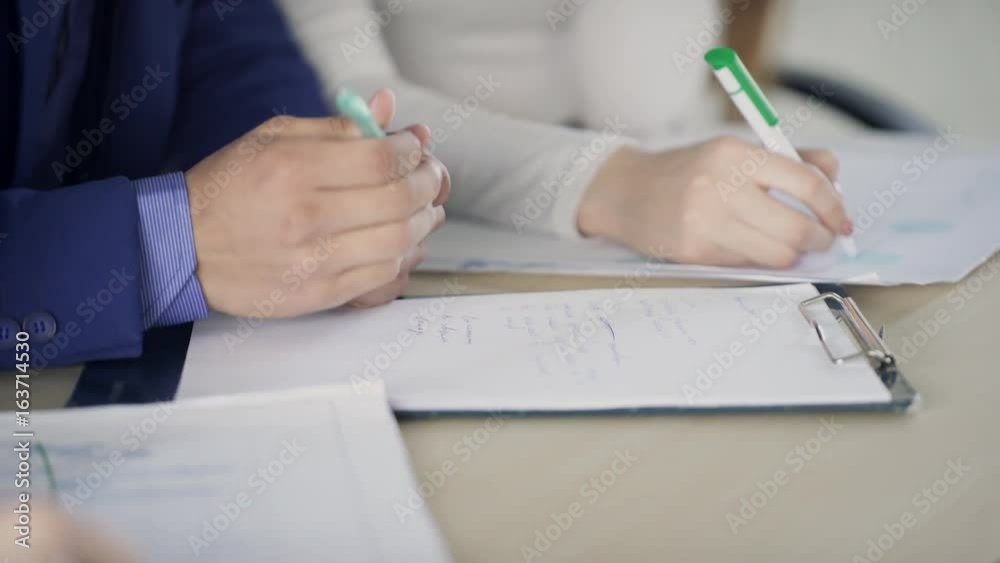 Male and female hands are writing on paper at the desk. The chief in a blue business suit shows the secretary, who keeps the record, about important issues that need to be reflected in the documents Male and female hands are writing on paper at the desk. The chief in a blue business suit shows the secretary, who keeps the record, about important issues that need to be reflected in the documents