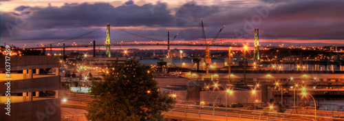  a nighttime view of a bridge with ocean and container pier, with street lamps lit and purple sky 