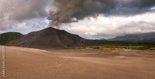 Mt Yasur volcano behind its rippled ash plain showing an eruption with smole billowing into the cloudy sky.
