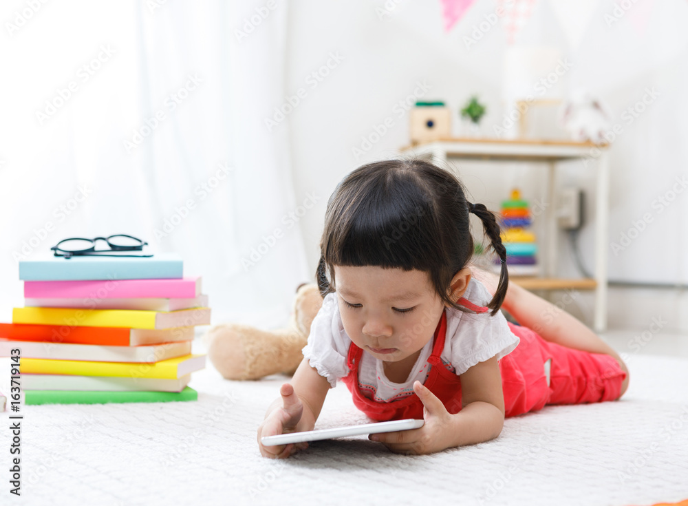 Naklejka premium Preschooler girl plaing tablet. Cute child reading with teddy bear. Little girl having fun indoors at home, kindergarten or